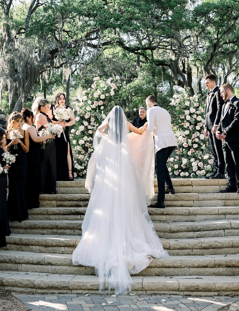 The bride and groom ascend wide stone stairs toward a large floral arch as the wedding party watches during the ceremony recessional.