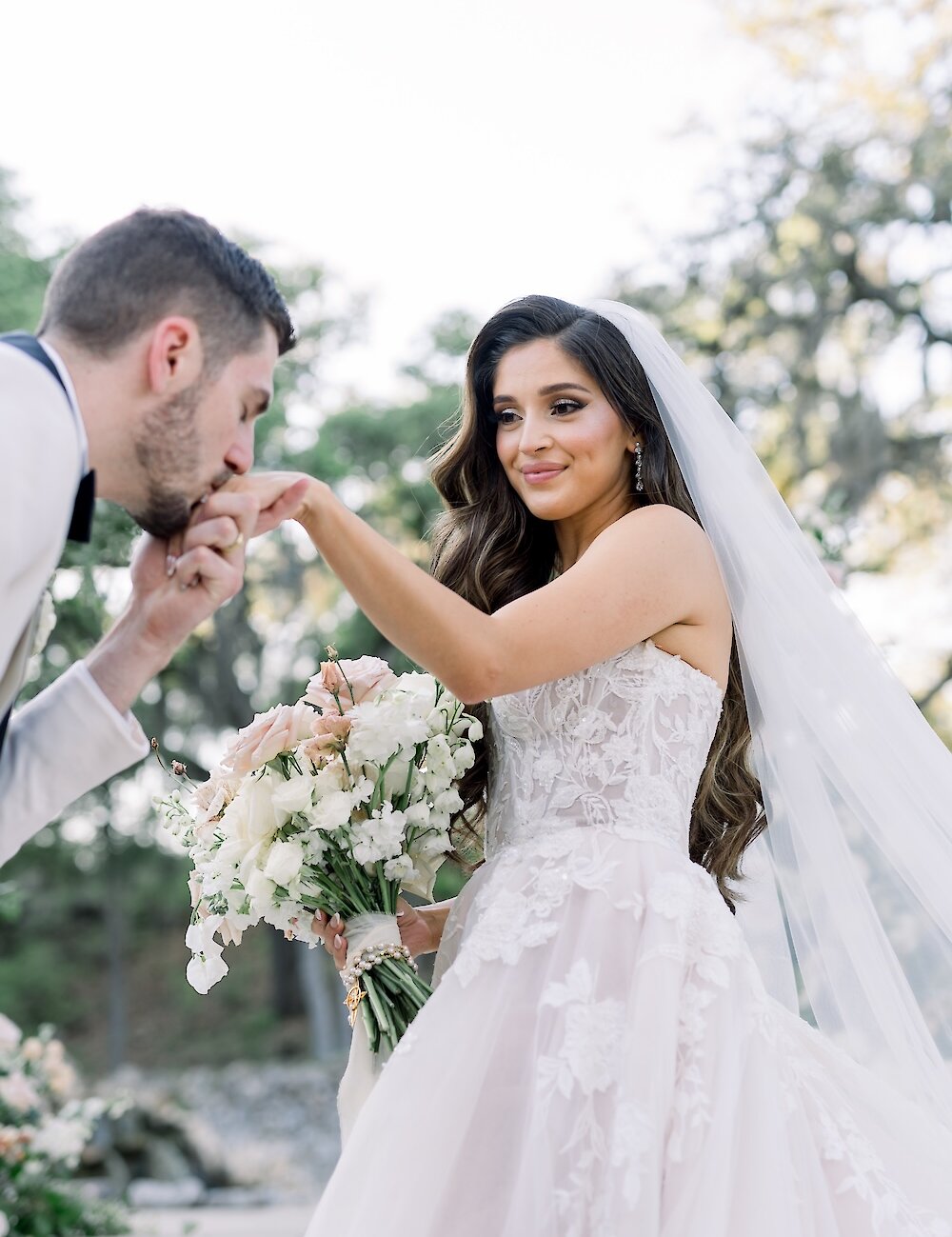 The groom in a white jacket gently kisses the bride's hand while she holds a peach and white bridal bouquet outdoors.