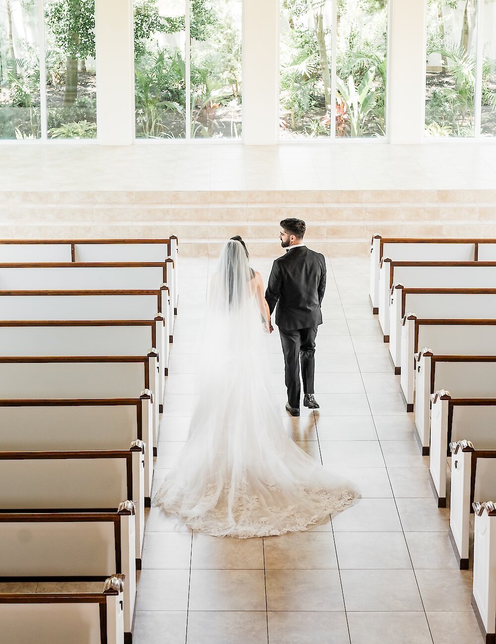 An overhead view of the bride and groom holding hands and walking away from the camera down the aisle toward a wall of bright windows.