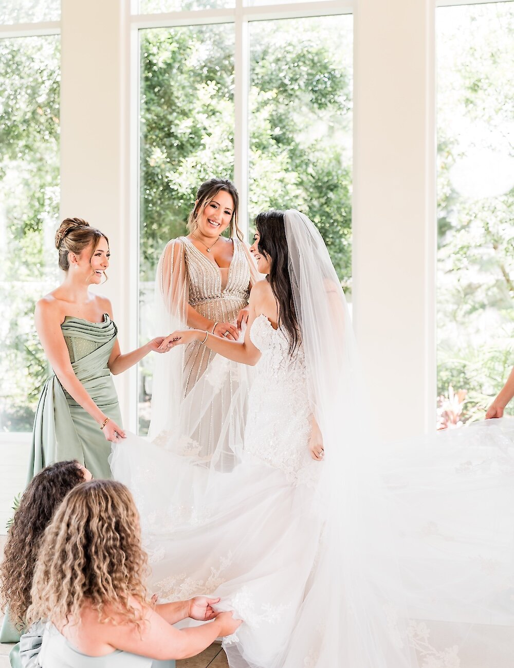 The smiling bride in her lace gown is assisted by her bridesmaids in light green and tan dresses near a wall of bright windows.