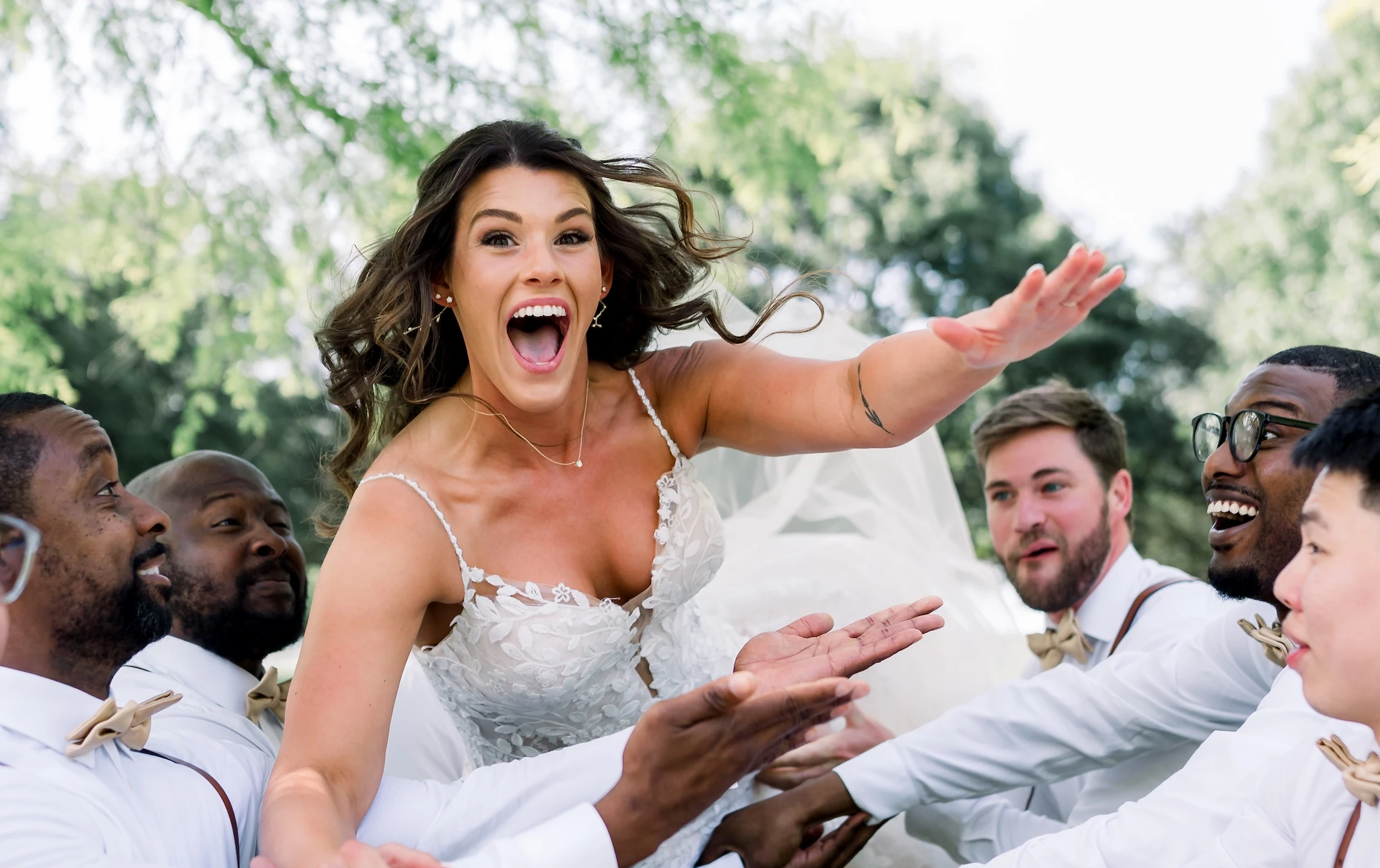 The ecstatic bride is being lifted and tossed playfully by the groomsmen dressed in white shirts and suspenders during an outdoor portrait session.