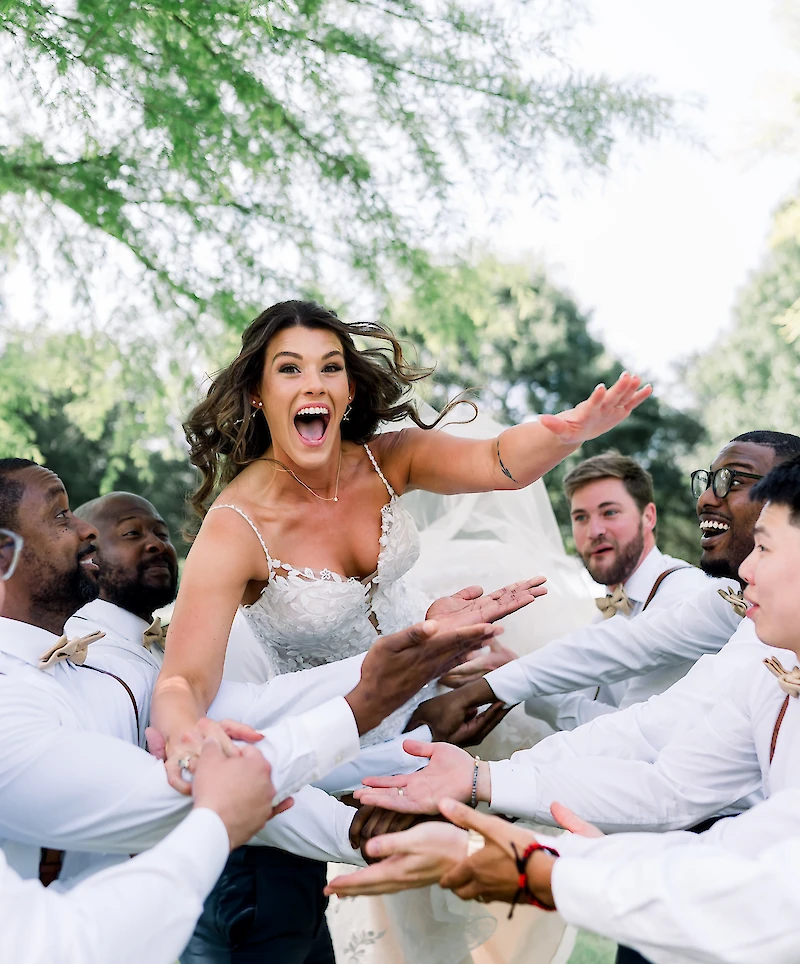 The ecstatic bride is being lifted and tossed playfully by the groomsmen dressed in white shirts and suspenders during an outdoor portrait session.