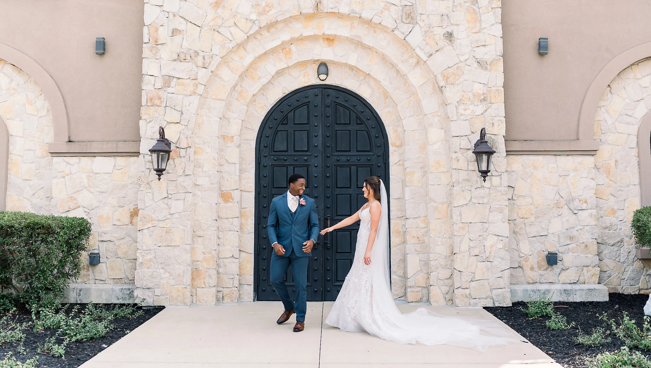 The bride in a lace gown smiles at the groom in a blue suit as they walk away from the arched black door of a stone church building.