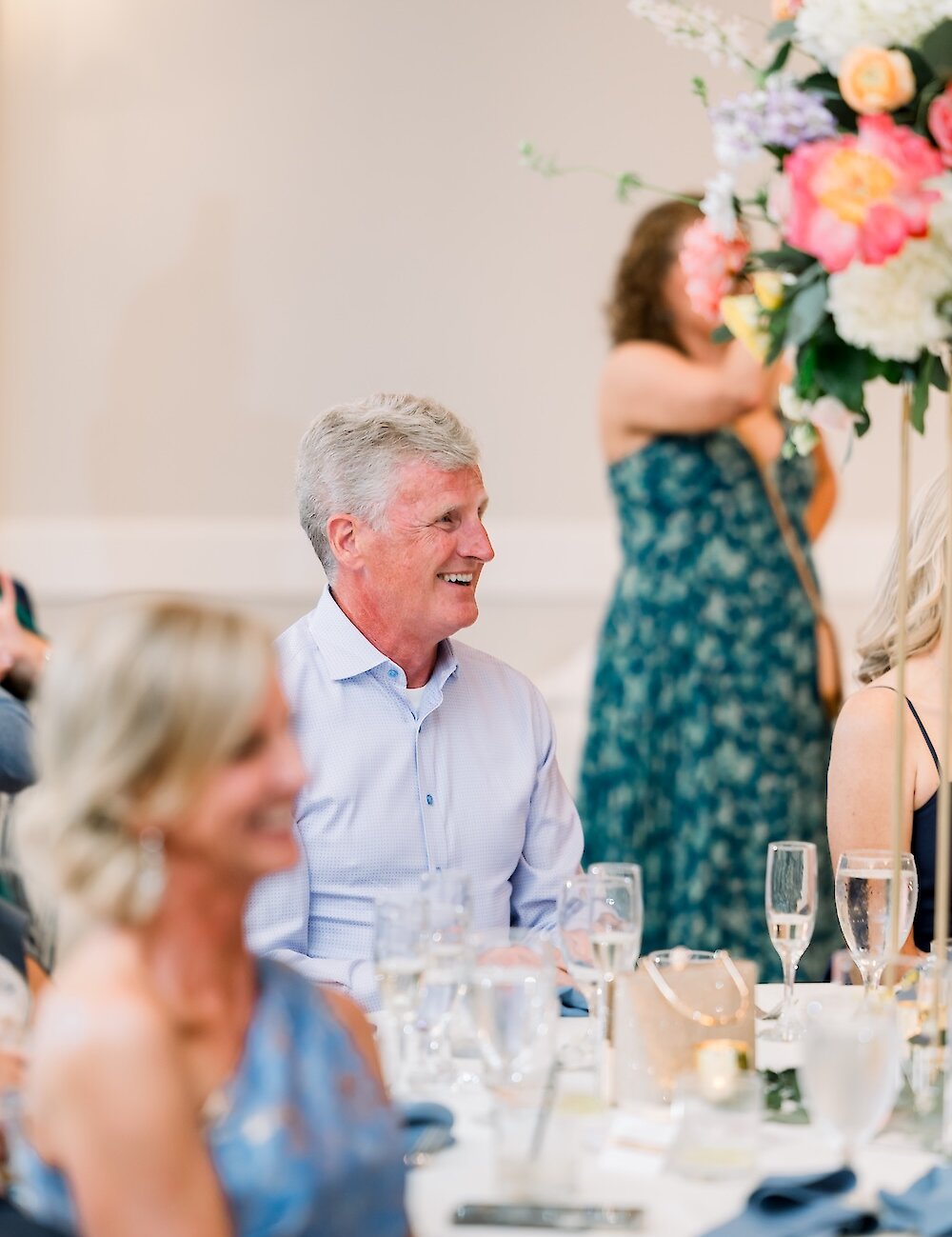 A smiling man wearing a light blue button-up shirt sits at a reception table decorated with tall floral arrangements and champagne glasses.