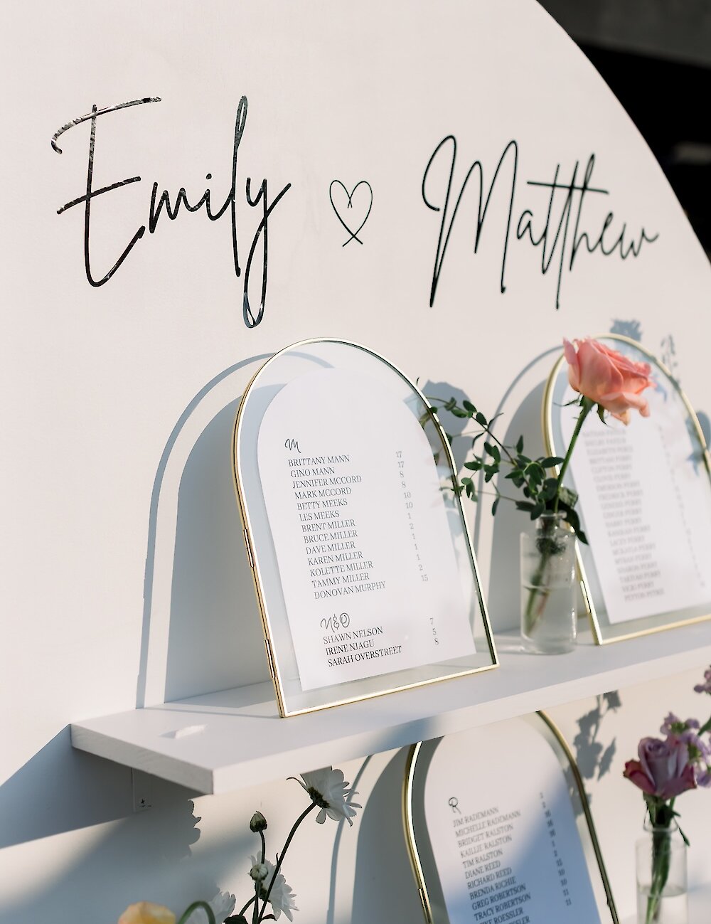 A modern arched white seating chart displaying the names Emily and Matthew, with individual guest lists in gold arched frames on a shelf.