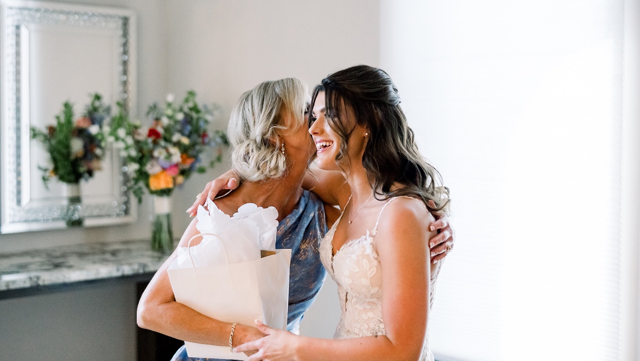 The smiling bride embraces her mother, who is wearing a blue and gold dress, while holding a white gift bag in the bridal suite.