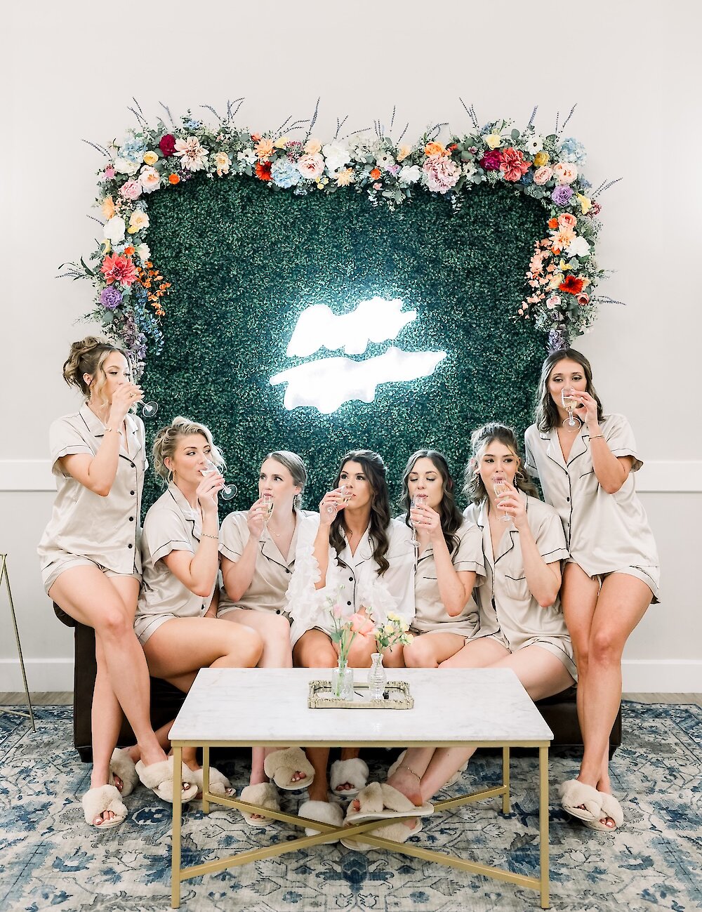 The bridal party in matching champagne pajamas cheers with champagne flutes in front of a green hedge wall decorated with a neon sign and colorful flowers.