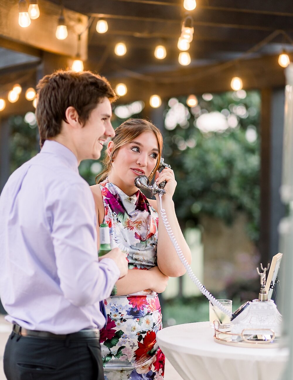A female guest in a floral dress uses a vintage telephone for an audio guest book as a male guest stands beside her under string lights at the outdoor reception.