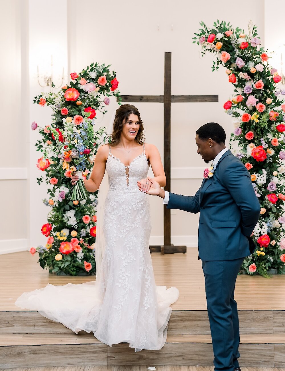 The bride and groom are holding hands, smiling at each other at the altar, which is framed by a wooden cross and two vibrant floral arches.