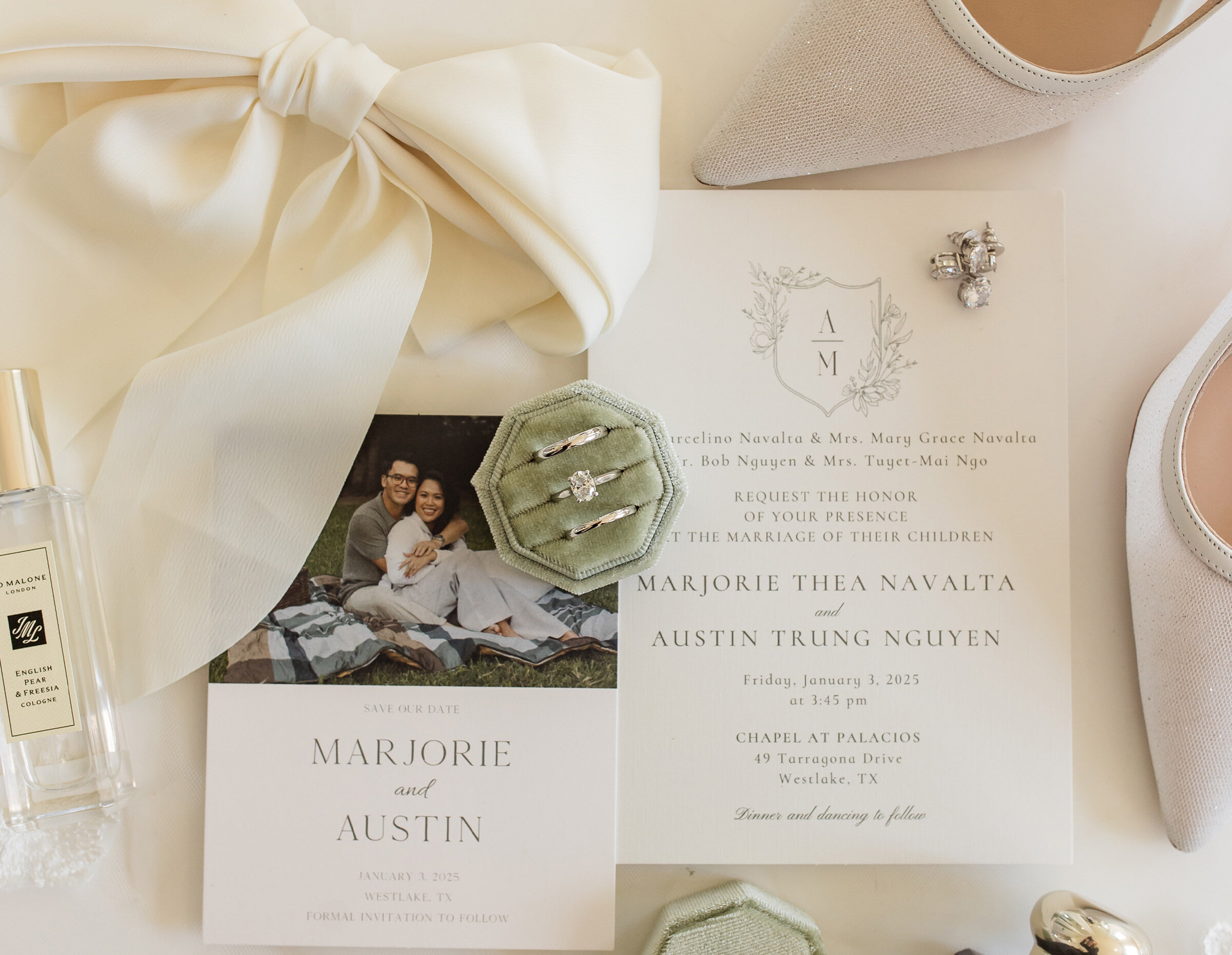 Flat lay of a classic wedding invitation suite with black and white typography, resting on a textured light background. Details include a black ribbon, pearl necklace, pearl earrings, wedding rings in velvet boxes, and a Jo Malone perfume bottle.