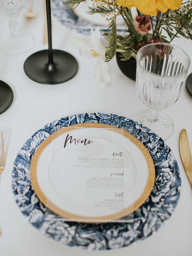 Close-up wedding reception table featuring a pink floral print tablecloth and a lush centerpiece of light pink, white, and green hydrangeas. Place settings include a hot pink arched menu card and gold votive candles.