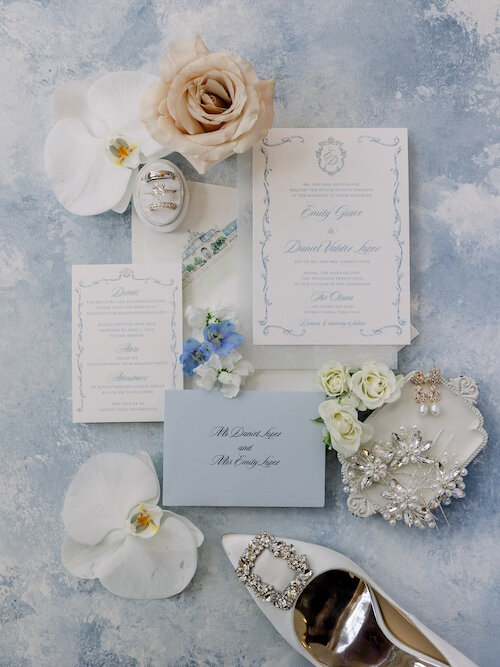 Elegant wedding invitation suite flat lay with white and light blue stationery, styled on a blue and white painted background. The arrangement includes wedding rings nestled in a box, white orchids, a nude rose, and a white satin shoe with a rhinestone buckle.