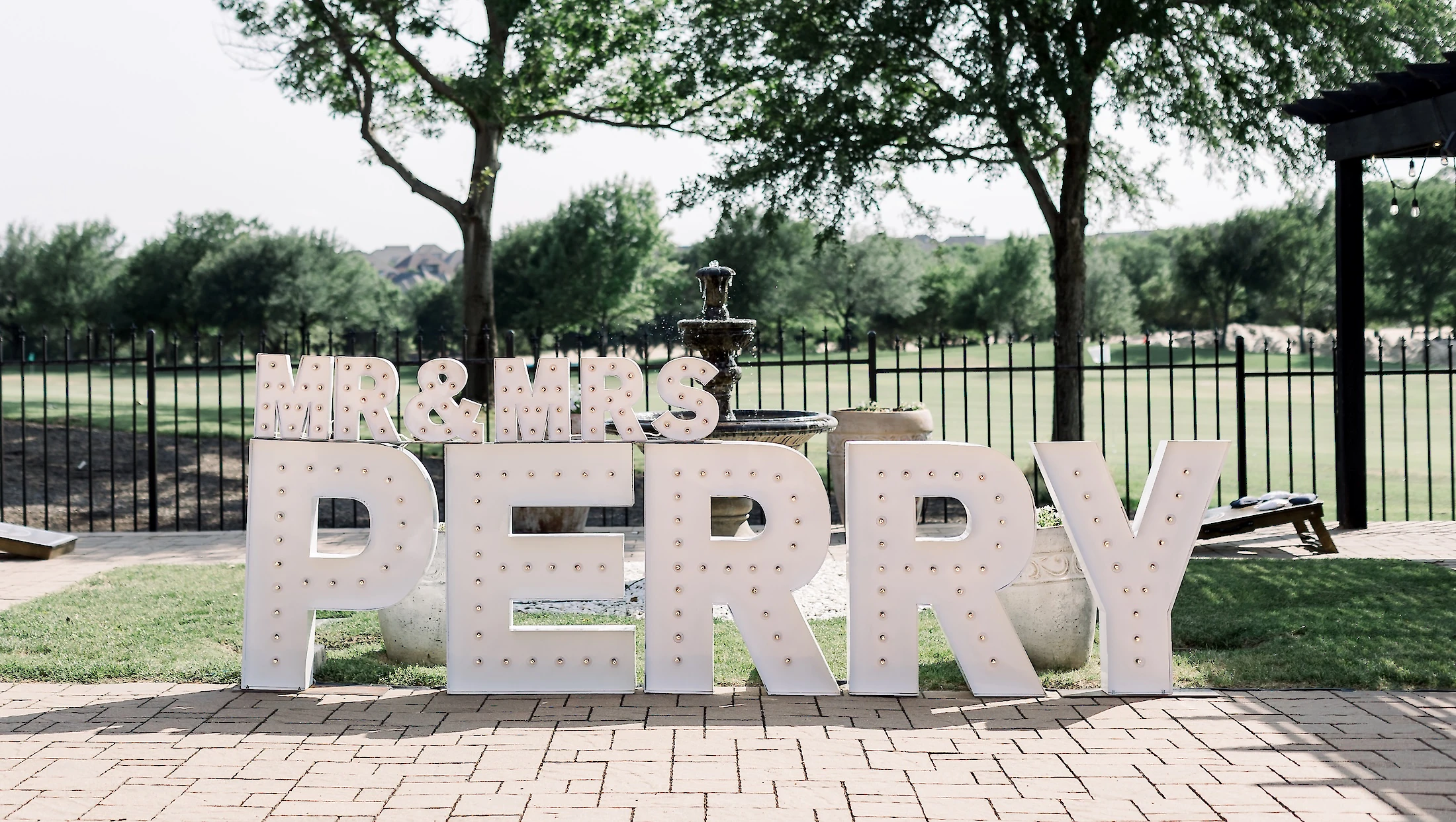Large outdoor marquee letters spelling 'MR & MRS PERRY' sit on a brick patio in front of a black fence, a fountain, and trees.