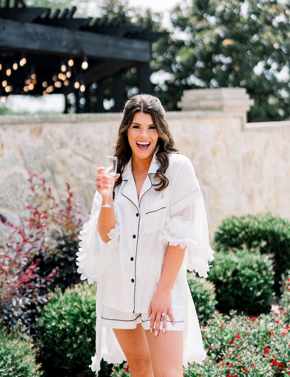 A happy bride poses outdoors in white pajamas with black piping and ruffles, holding a champagne flute, surrounded by green shrubs and a stone wall.