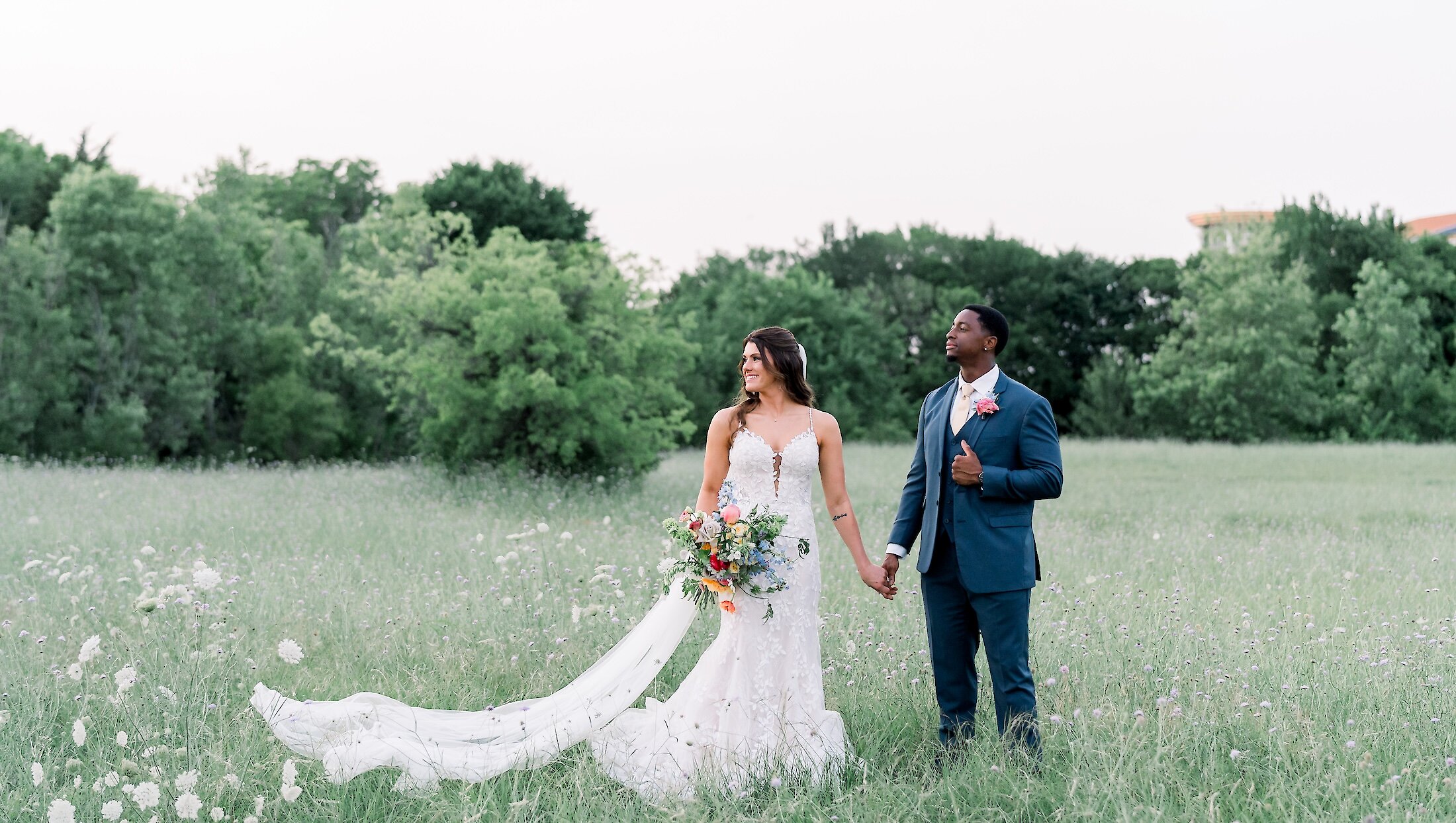 The bride and groom stand holding hands in a grassy summer field, with the groom in a blue suit and the bride's lace train flowing behind her.