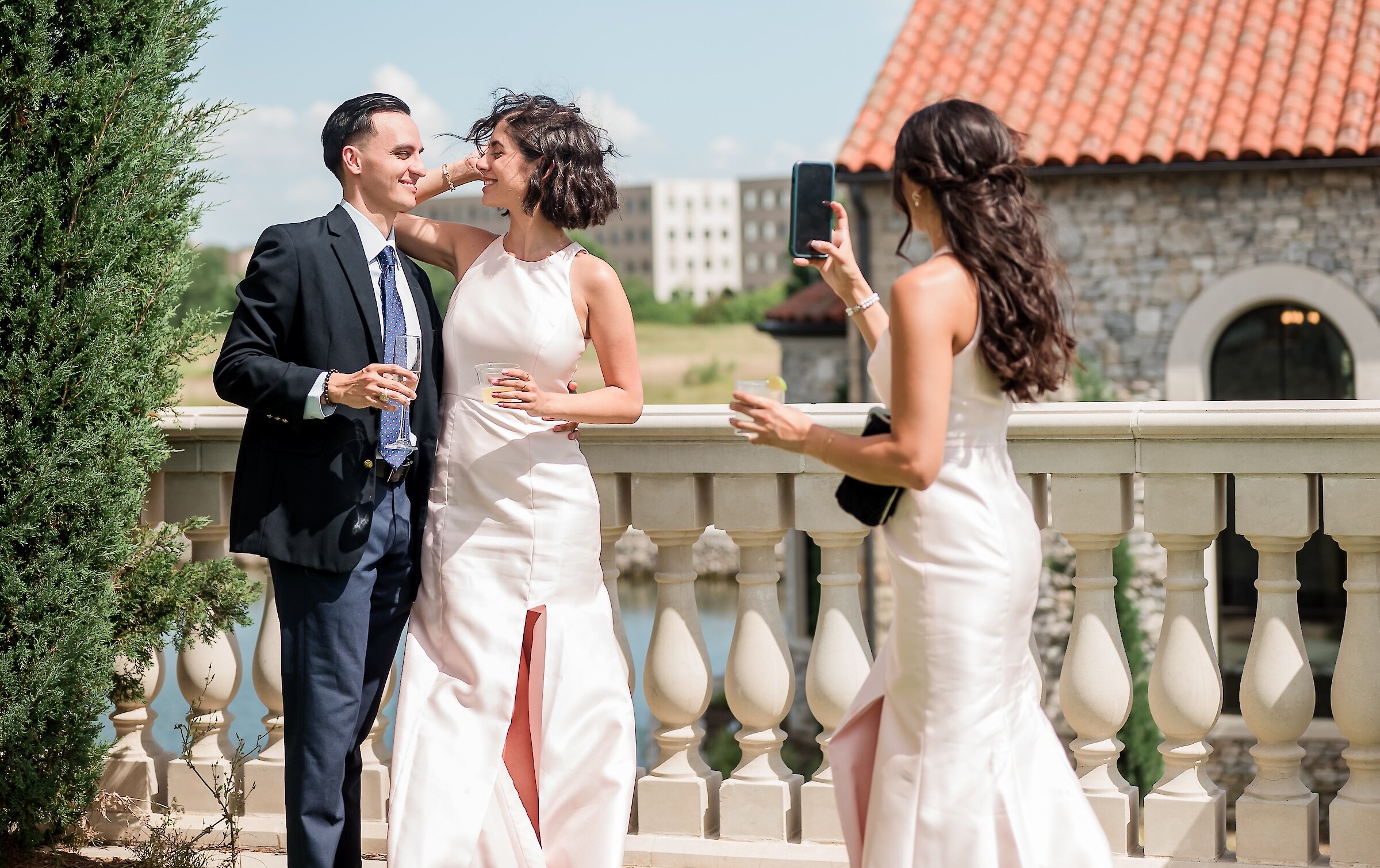 A group of wedding guests taking a picture on a sunny outdoor patio with turf, a balustrade, and a stone building with a terracotta roof.