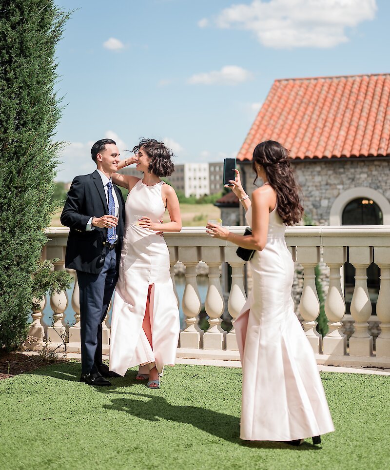 A group of wedding guests taking a picture on a sunny outdoor patio with turf, a balustrade, and a stone building with a terracotta roof.