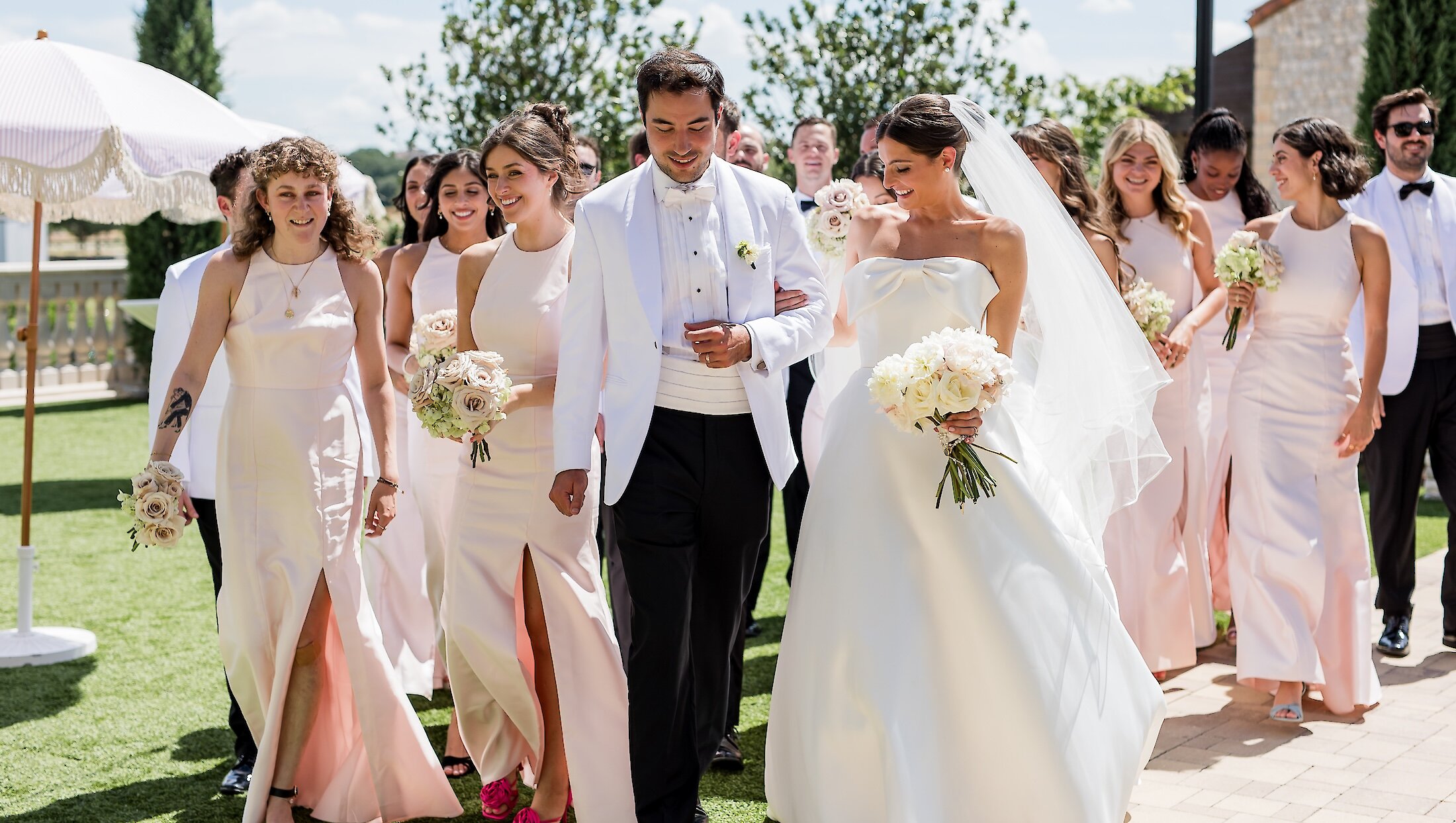 The smiling bride and groom walk down a path surrounded by their wedding party, with bridesmaids in blush dresses and groomsmen in white jackets.