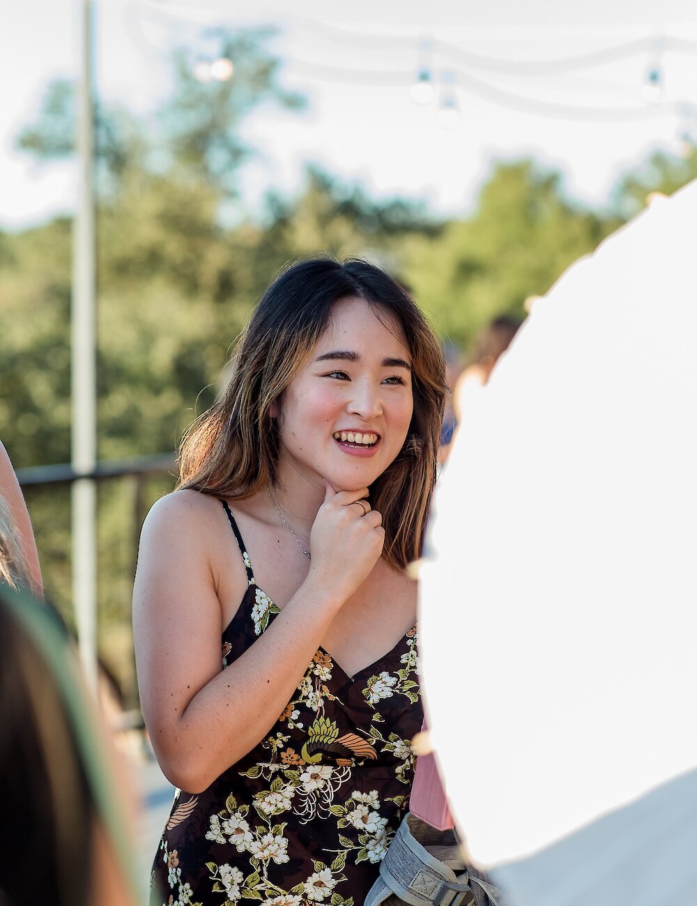 A smiling wedding guest in a dark floral print sundress holds her chin, talking to someone under string lights outdoors on a sunny day.