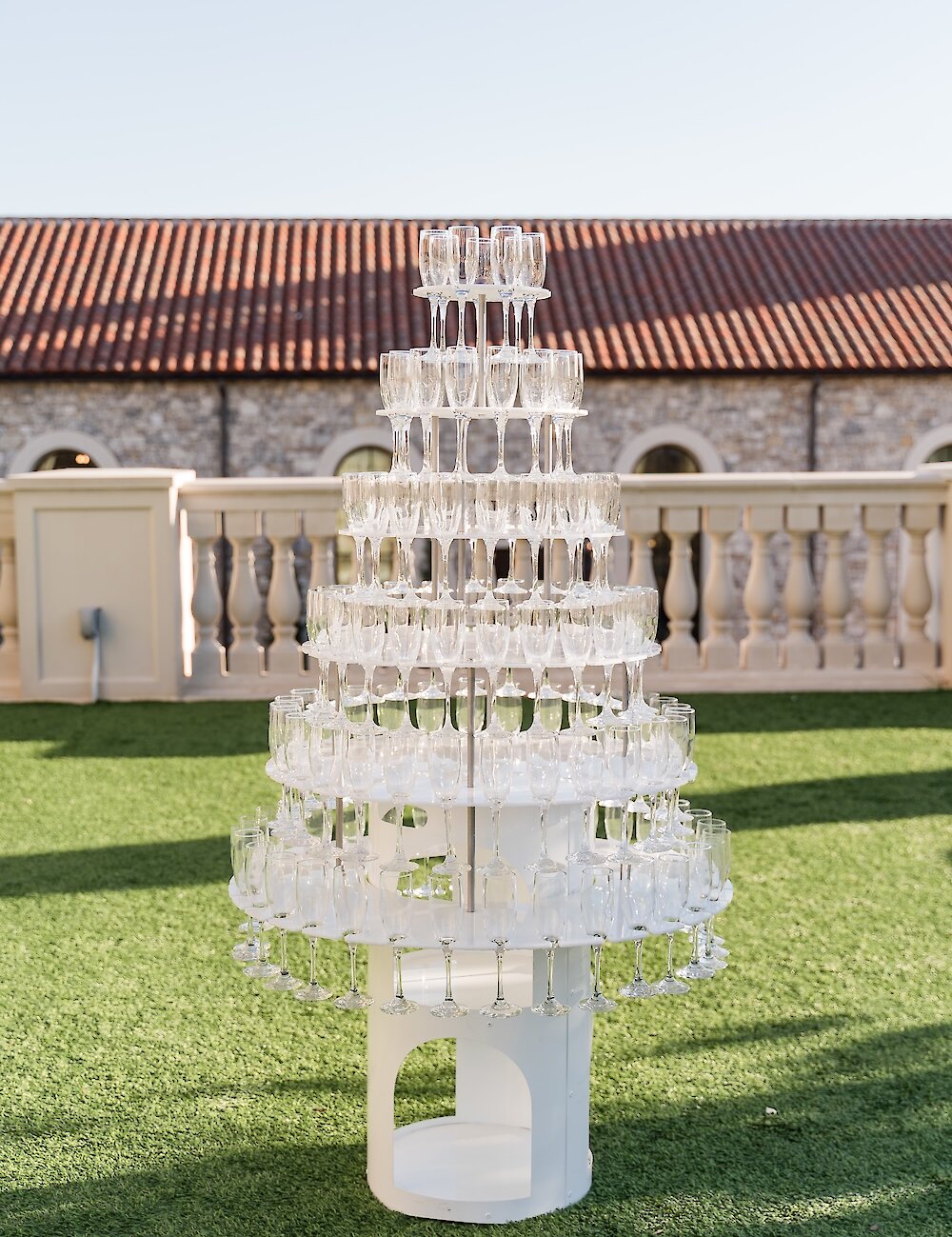 A multi-tiered champagne glass tower display stands on artificial turf outdoors against a white balustrade and a stone building with a red tile roof.