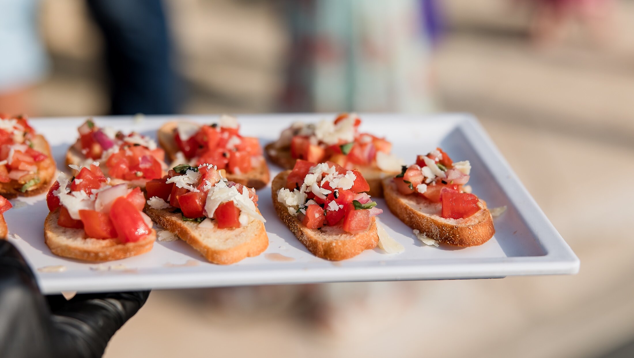 A caterer wearing black gloves holds a white tray of fresh bruschetta appetizers topped with diced tomatoes, red onion, and grated cheese.