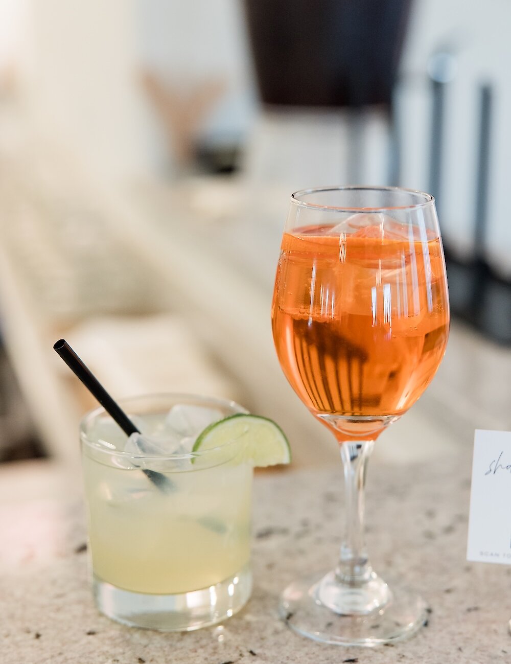 Two wedding cocktails, one orange aperol spritz in a wine glass and a pale margarita with a lime wedge, rest on a light granite bar top.