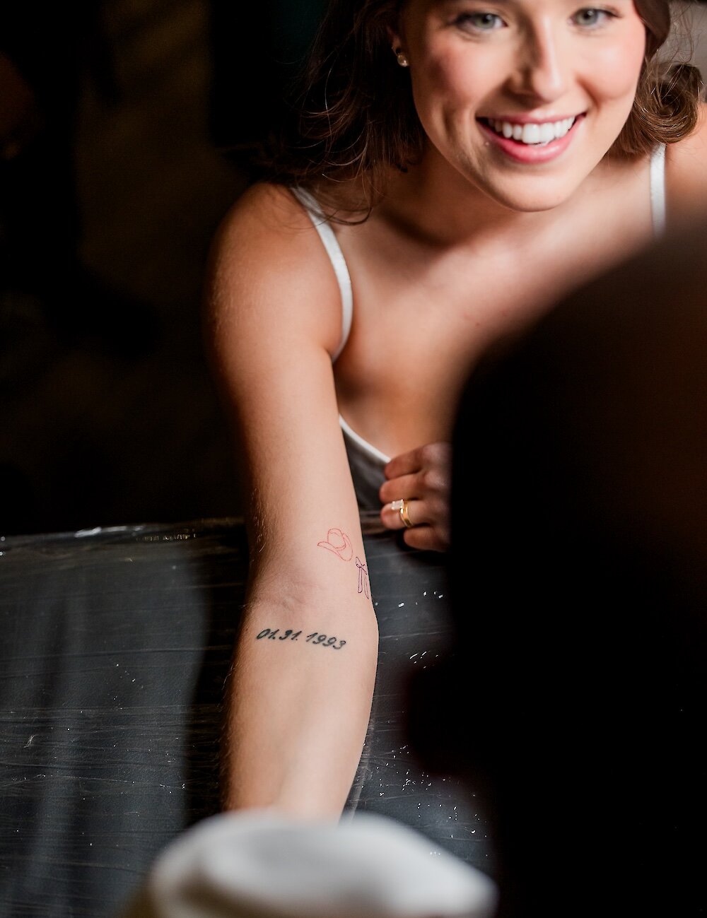 The smiling bride shows her forearm tattoo of the date "04.31.1993" and a small red cowboy hat drawing to someone in the foreground.