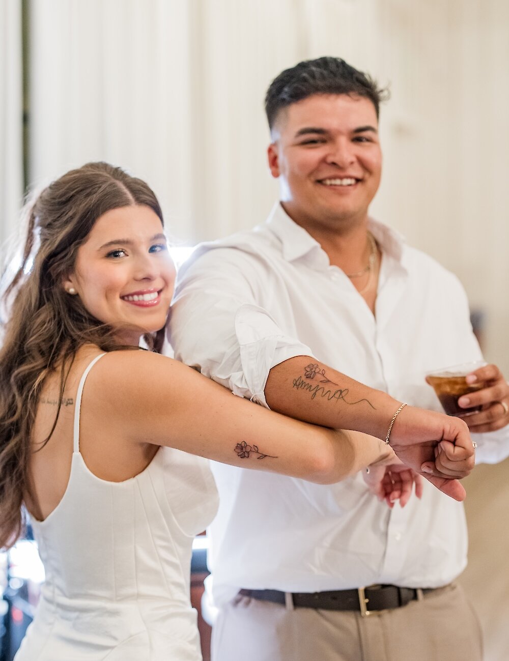 The bride and groom smile while holding up their forearms to display small, matching floral tattoos to the camera.