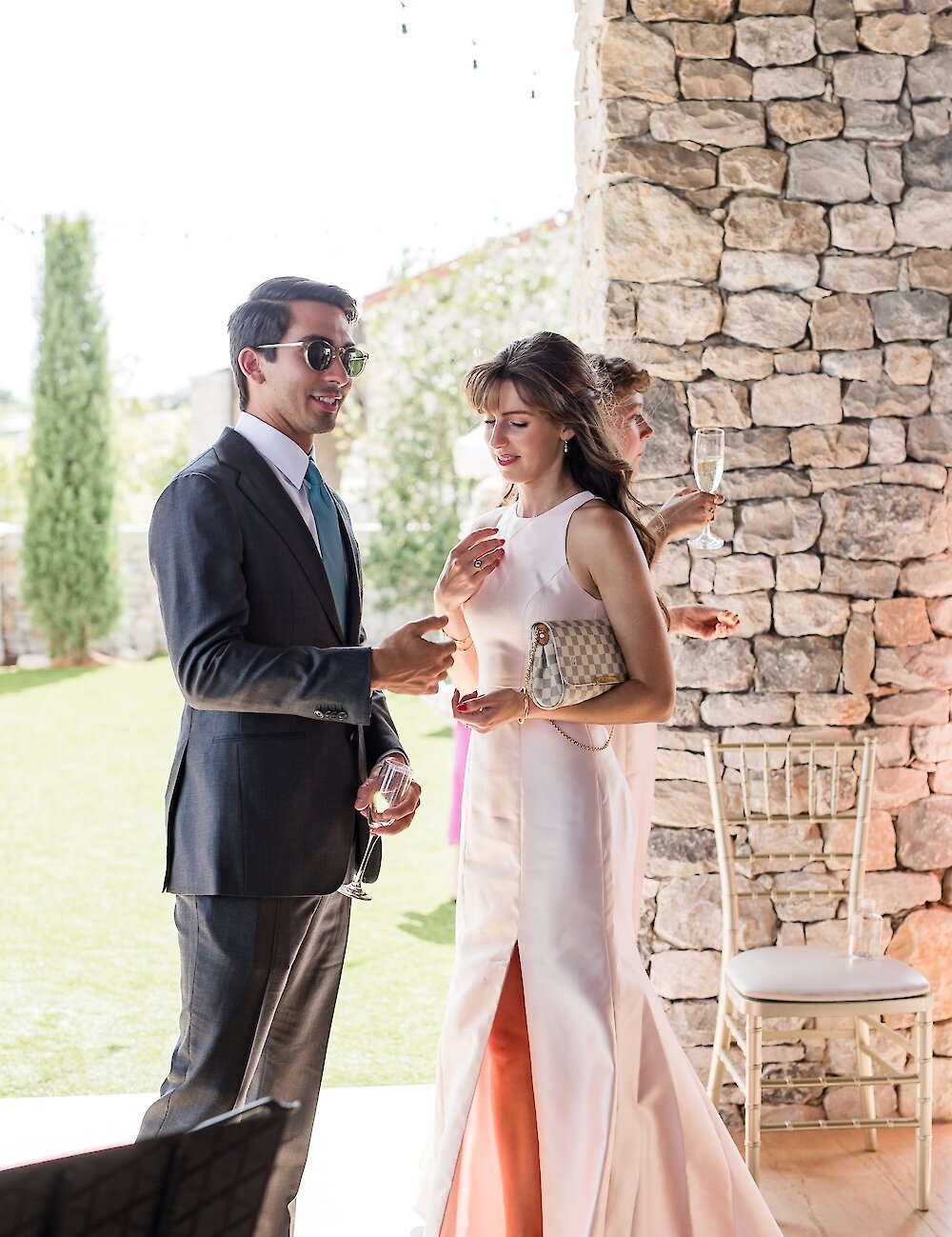 A male guest in a dark suit talks to a woman in a pale pink high-slit dress while holding a champagne glass under a covered patio with a stone wall.
