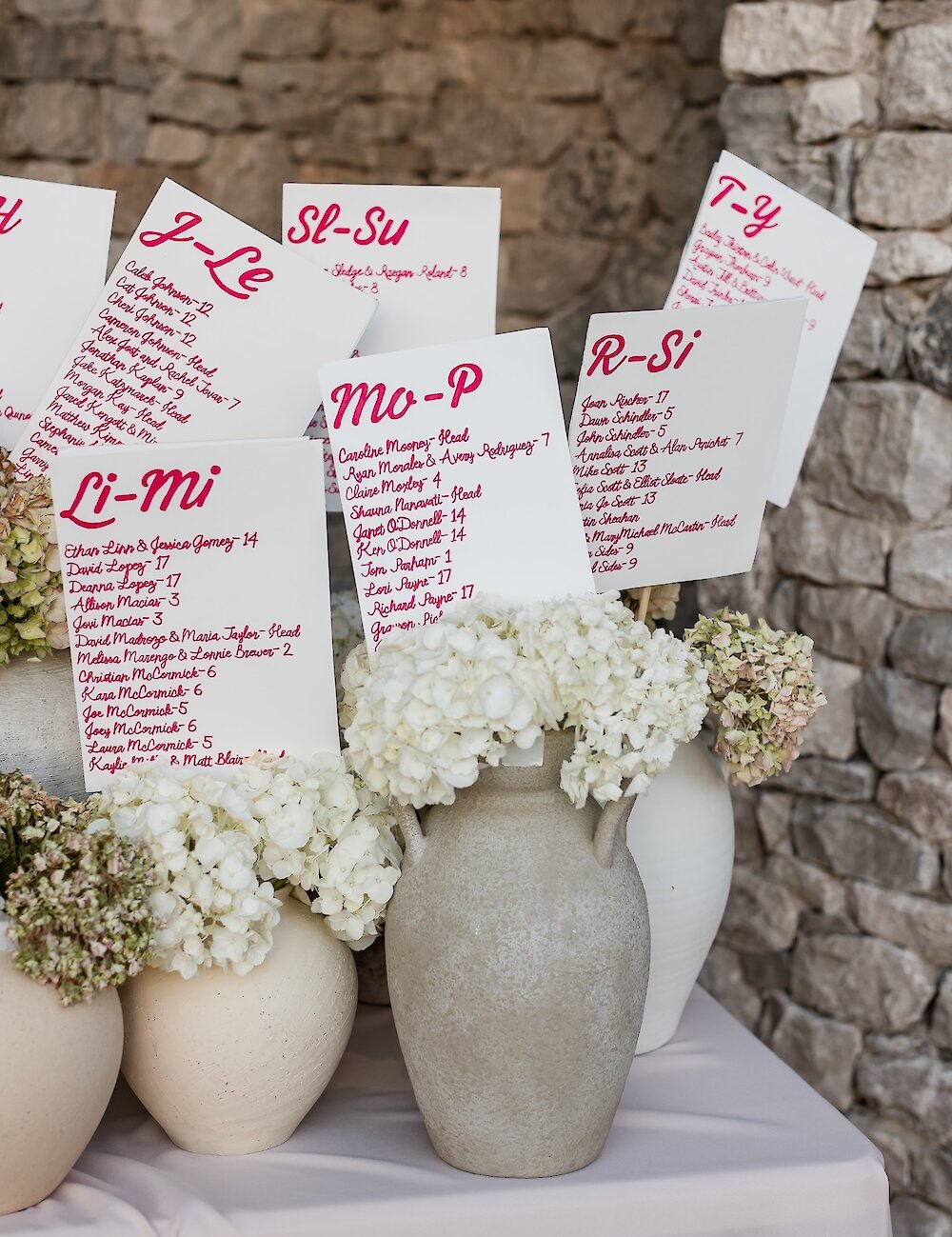 Individual white seating chart cards with pink lettering are displayed in vases of white hydrangeas against a rustic stone wall background.