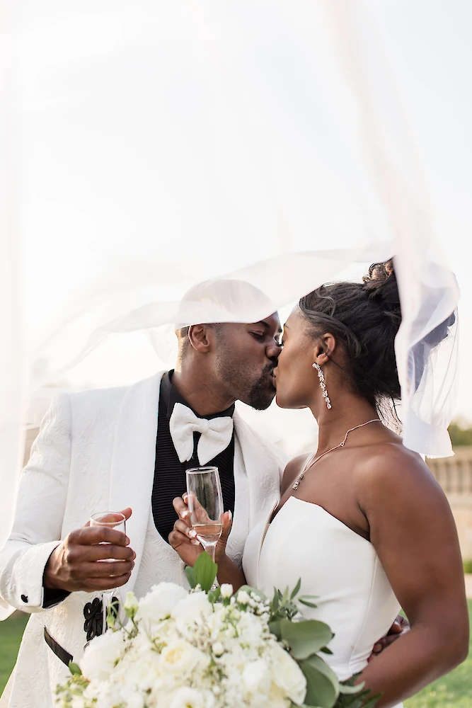 The bride and groom share a kiss under her sheer veil while holding champagne flutes and the bride's white floral bouquet.