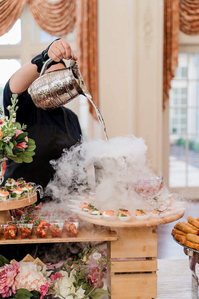 A server pours liquid onto dry ice, creating a cloud of smoke around an elegant wedding appetizer display featuring small bites.