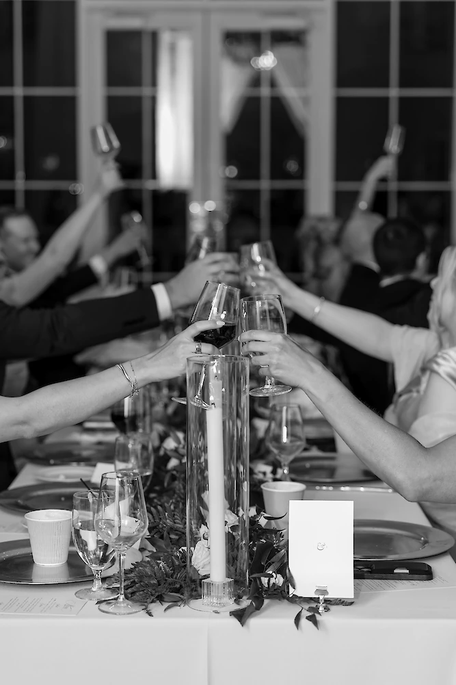 A black and white photo of wedding guests raising wine glasses for a toast over a dinner table with tall candle centerpieces.