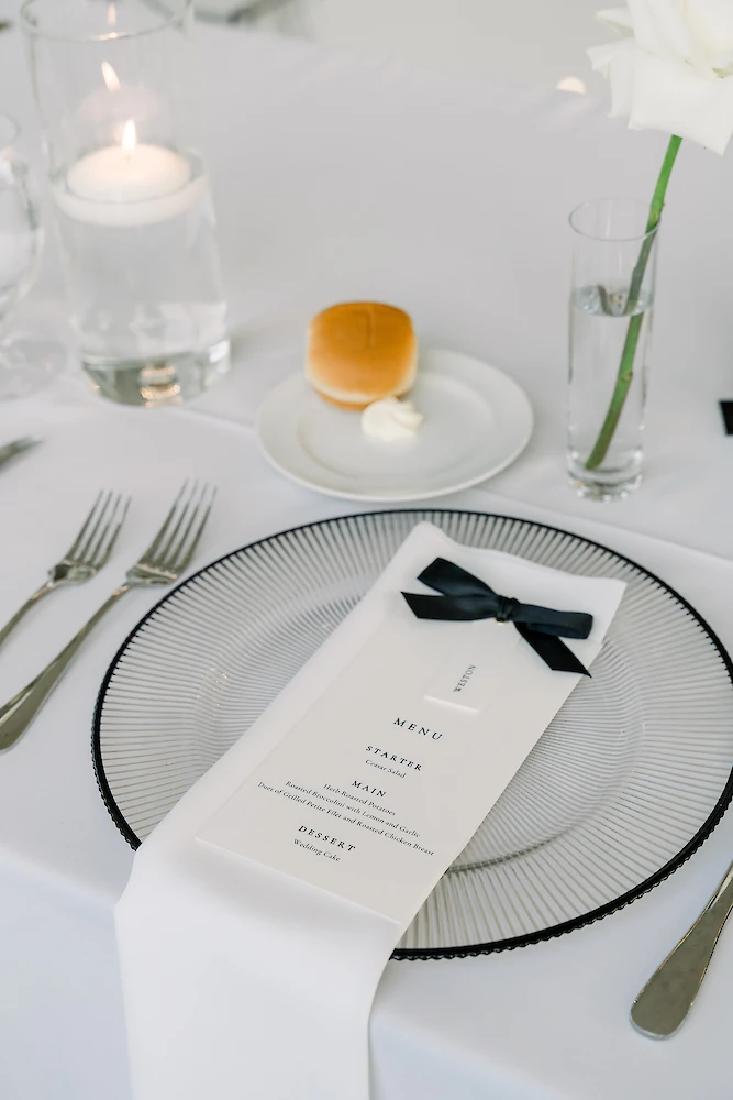An elegant wedding place setting featuring a ribbed glass charger plate, a menu card with a black ribbon, and a roll with butter.