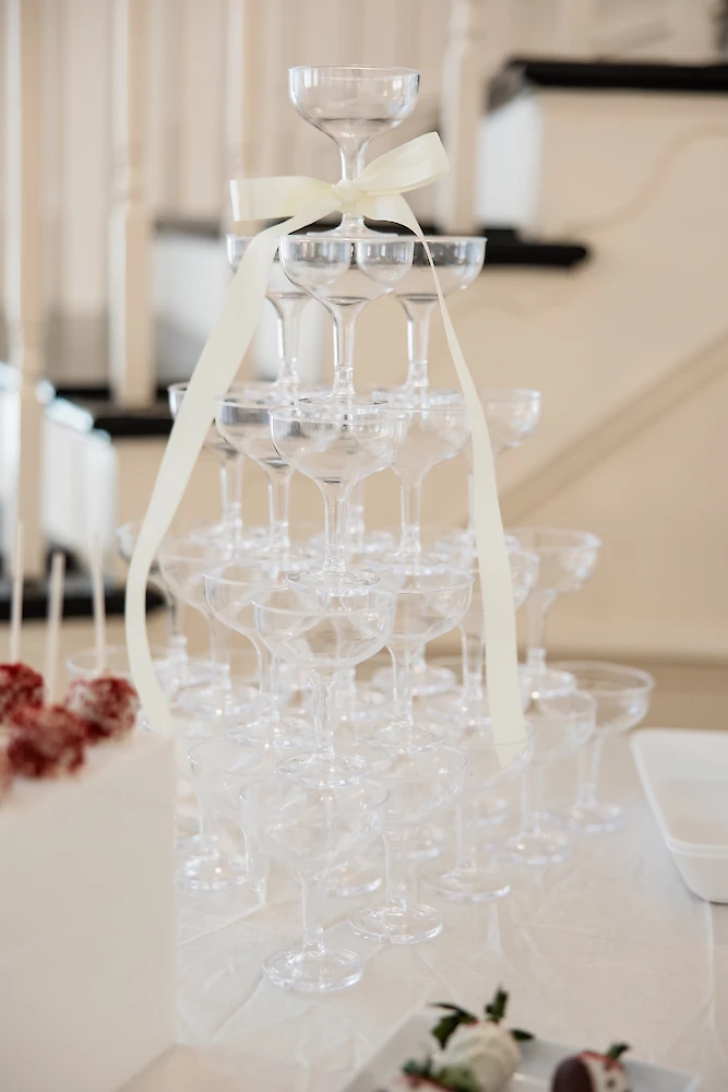 A tiered tower of empty champagne saucer glasses topped with a delicate cream ribbon, set up for a wedding celebration.
