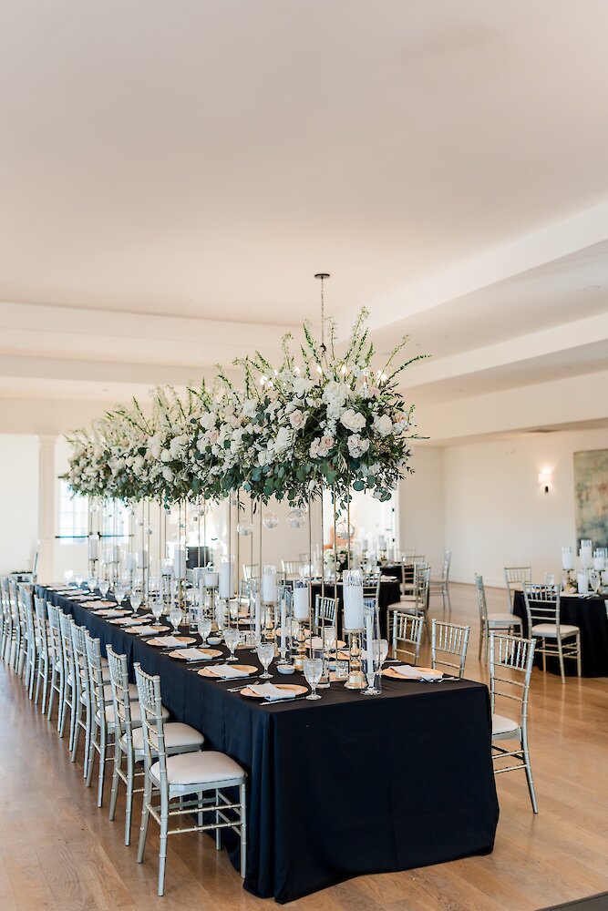 A long wedding reception table with a black linen and gold settings, featuring dramatic white and green elevated floral chandeliers.