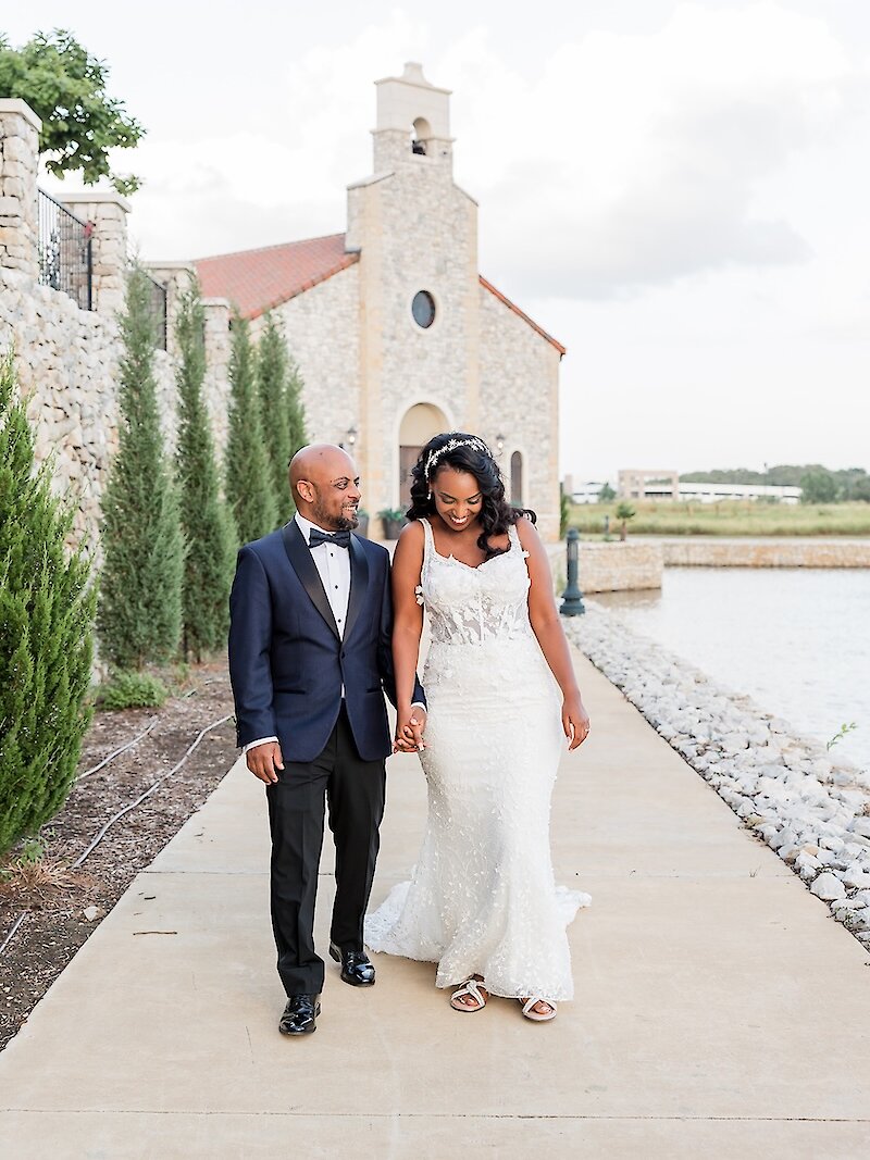 Candid wedding portrait of an African couple walking hand-in-hand along a waterside path, smiling at each other. The groom wears a navy blue tuxedo, and the bride wears a beaded, fitted gown with a long veil, with a stone chapel visible in the background.
