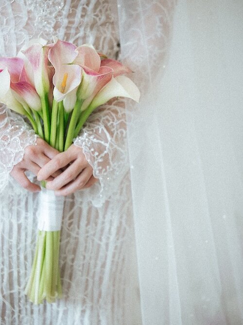 Close-up detail photo of a bride holding a tightly wrapped bouquet of pink and white calla lilies. The shot highlights the intricate texture of her long-sleeved lace wedding gown and sheer veil.