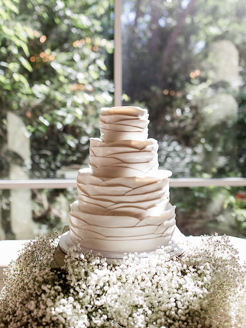Five-tier modern white wedding cake with sculpted fondant ruffles displayed on a table surrounded by a bed of baby’s breath flowers, set in front of a large window with natural greenery in the background.
