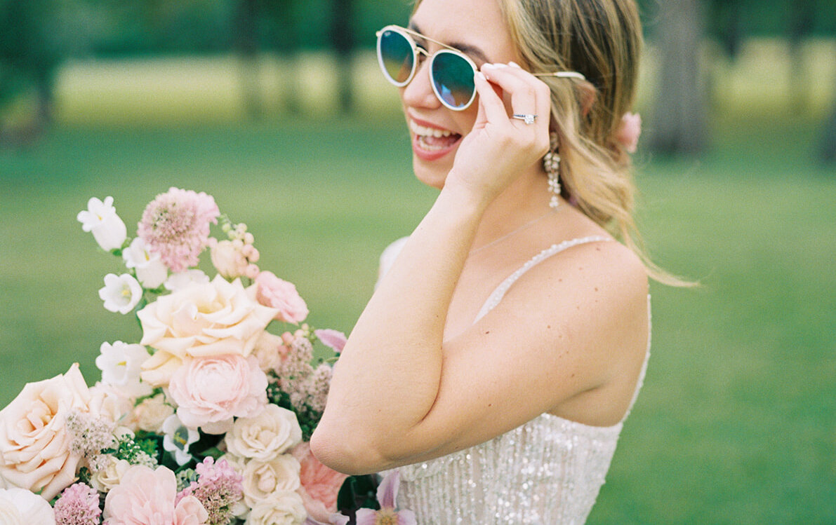 The joyful bride laughs and adjusts her mirrored sunglasses while holding a large bouquet of pink and peach roses on a sunny lawn.