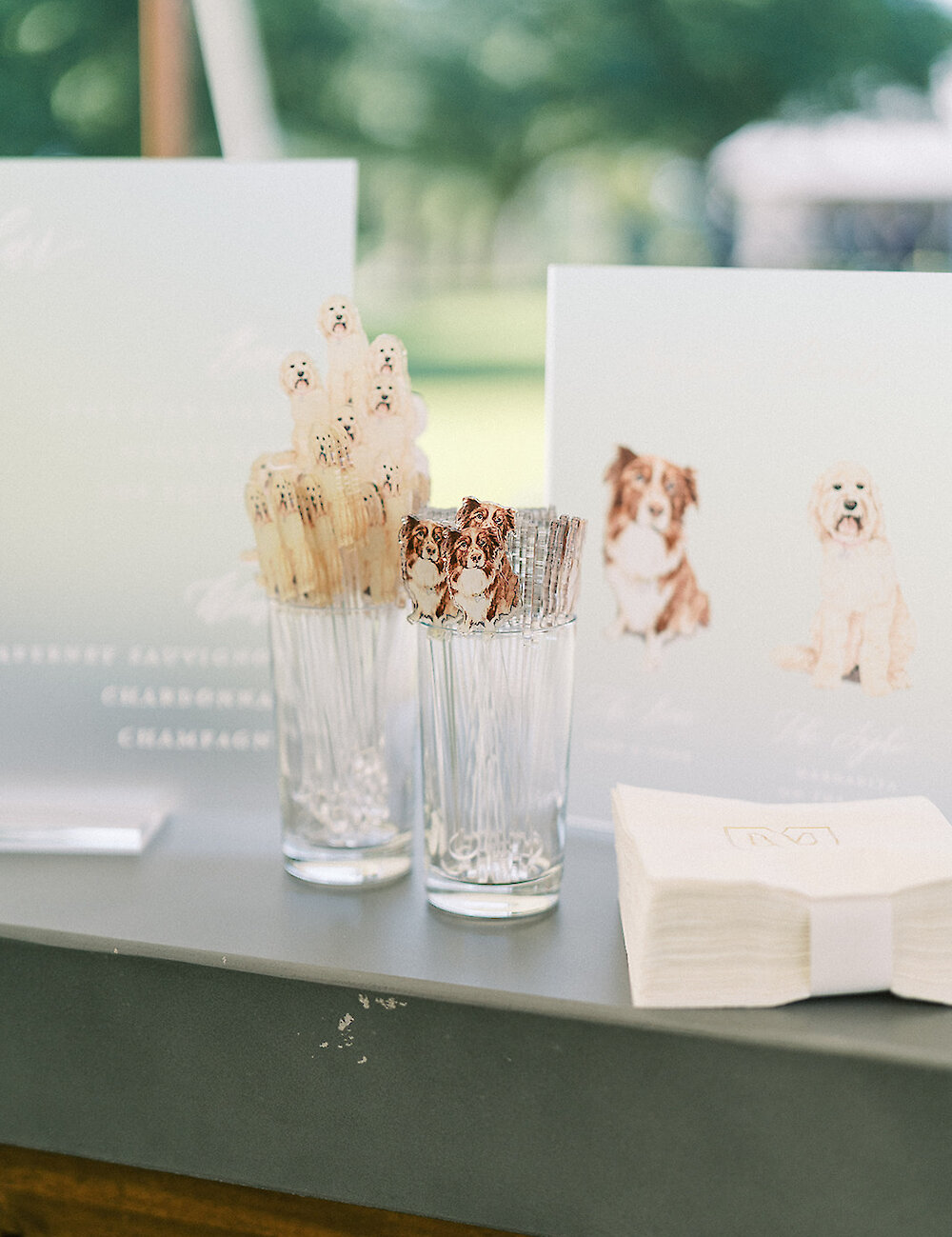 Clear glasses hold custom cocktail stirrers shaped like the couple's dogs, set next to a bar menu featuring dog illustrations and white cocktail napkins.