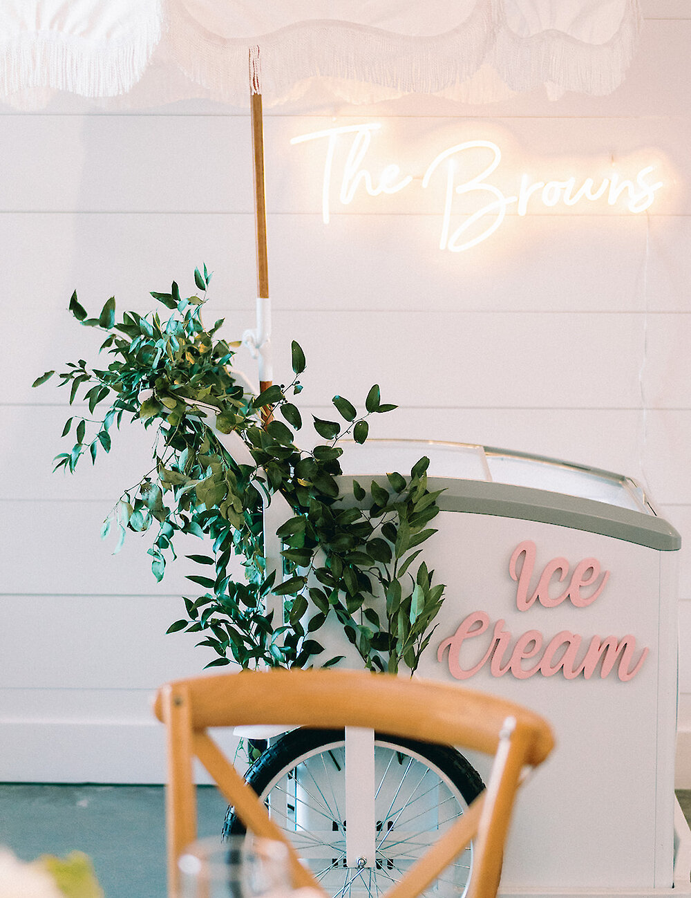 A white ice cream cart with a fringed umbrella and a neon sign that reads 'The Browns' is decorated with green foliage against a white shiplap wall.