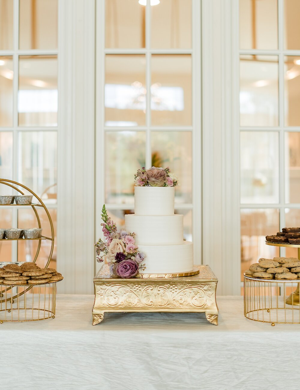 A three-tier white wedding cake decorated with mauve and purple roses sits on a gold stand, surrounded by cookies and other treats on a dessert table.