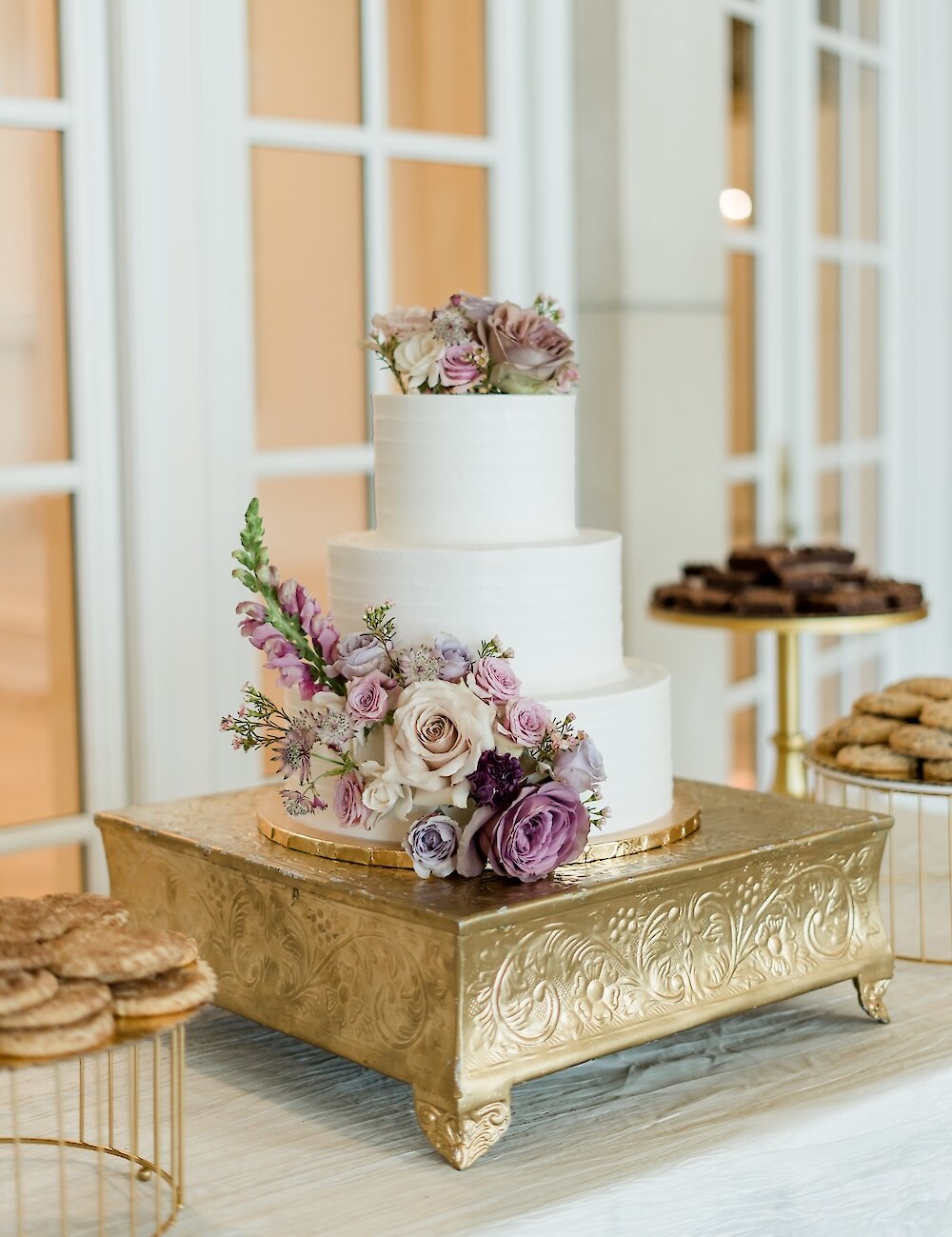 A three-tiered white wedding cake sits on a textured gold cake stand, lavishly decorated with mauve, purple, and pink roses and snapdragons.