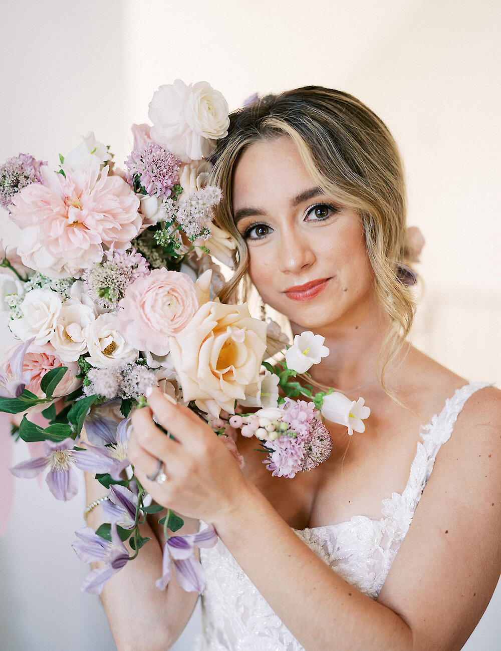 Close-up portrait of the smiling bride, holding a large pastel bouquet of pink dahlias, peach roses, and purple clematis, wearing a lace-strap gown.