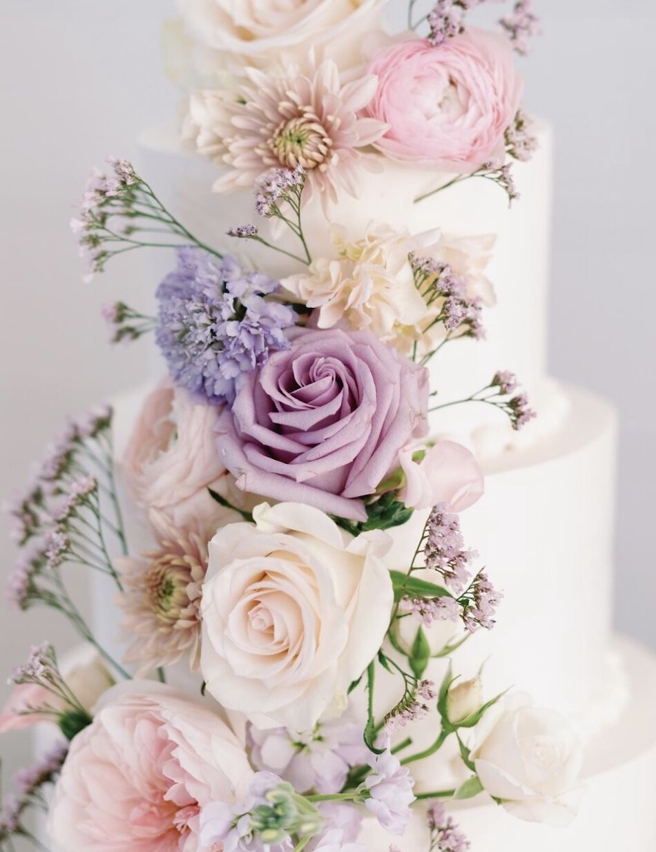 A close-up of a tiered white wedding cake decorated with a dramatic cascade of flowers, including lavender roses, pink ranunculus, and mauve accents.