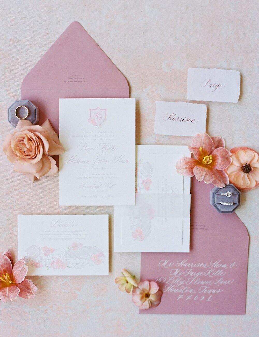A wedding invitation flat lay featuring blush and mauve envelopes, light pink watercolor monograms, a diamond ring, and peach ranunculus flowers.