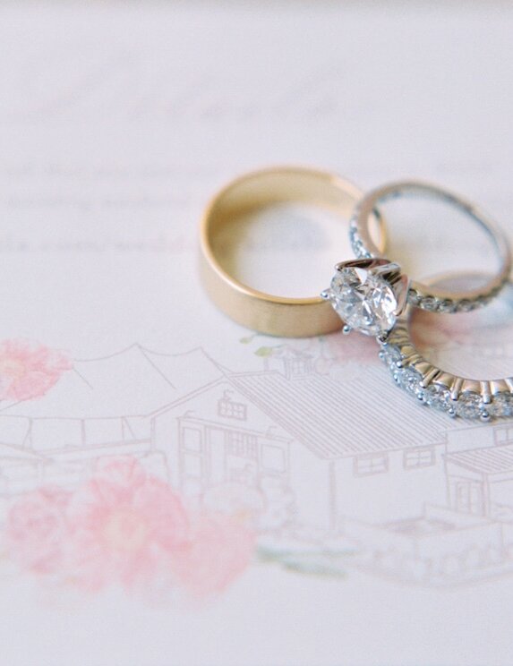 A close-up of wedding rings resting on the invitation, which features a faint, soft pink illustration of the wedding venue.