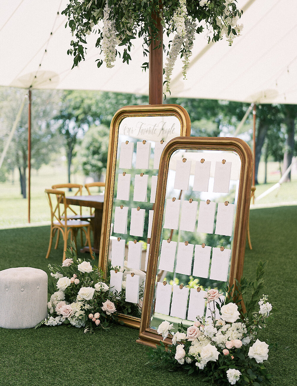 Two ornate gold-framed mirror seating charts stand on turf under a white tent, decorated with white and blush roses and hanging greenery.