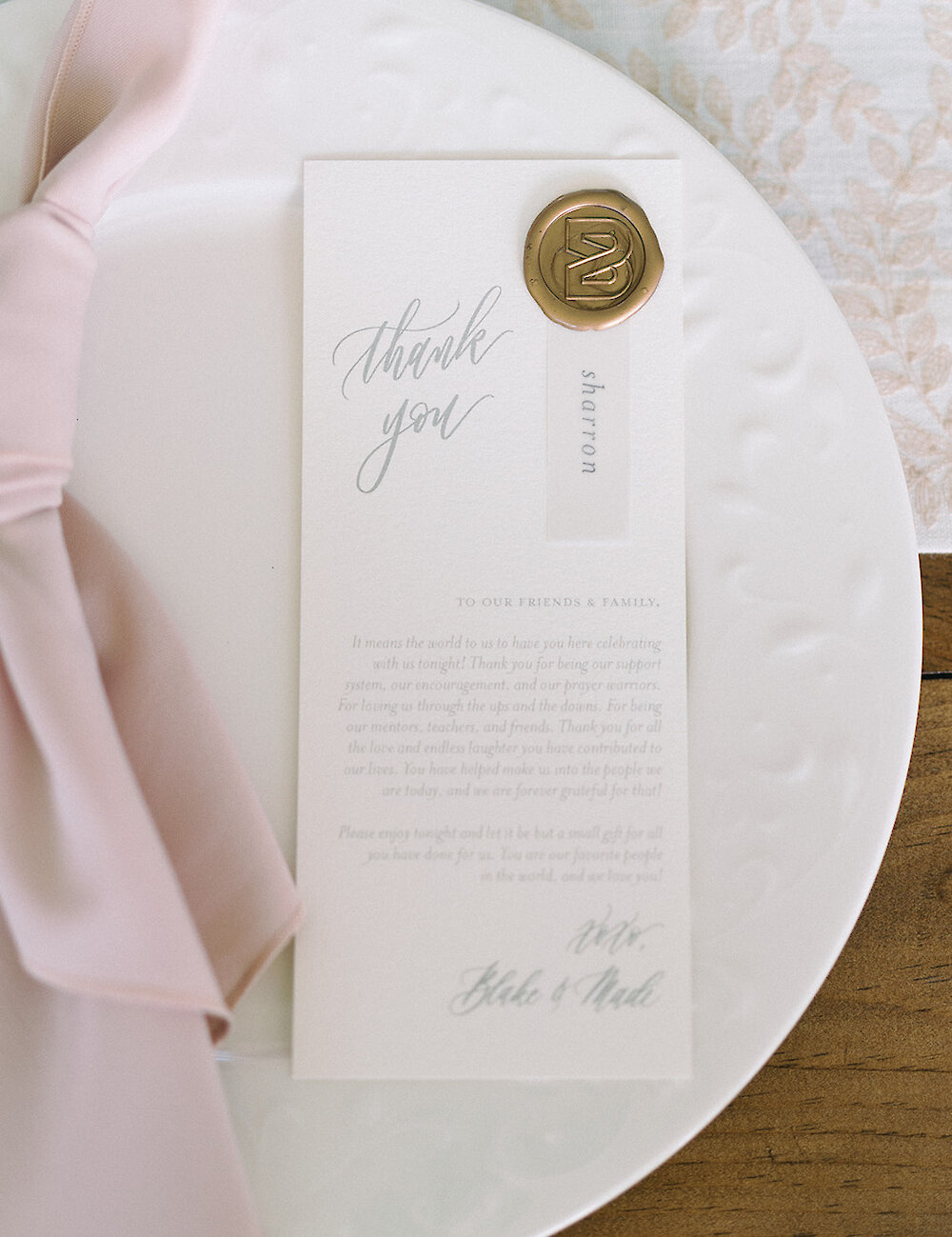 A wedding thank you card with a gold wax seal and guest name sits on a white plate, next to a blush pink napkin at a reception place setting.